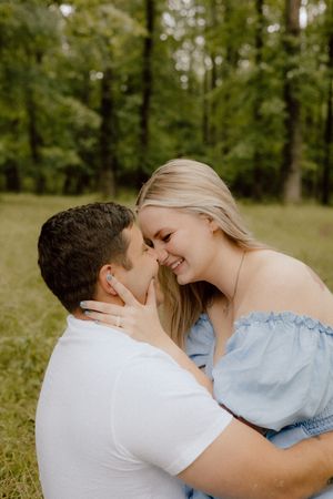 A romantic outdoor couple portrait shows two people sharing a tender moment while sitting in a green forest setting.