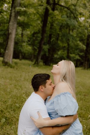 A romantic couple embraces on grass in a wooded area, with the person wearing a light blue dress leaning back.