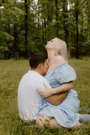 A couple shares an intimate moment while sitting together in a grassy meadow surrounded by trees on a summer day.