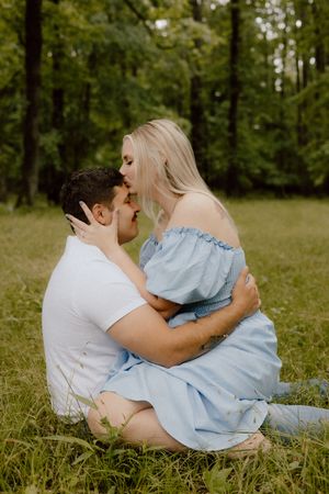 A couple shares an intimate moment while sitting together on grass in a wooded outdoor setting.