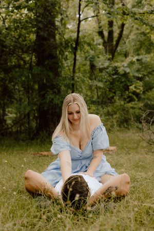 A person in a blue dress sits in a grassy meadow surrounded by trees on a sunny summer day.