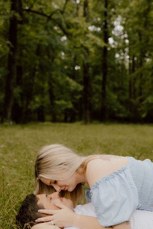 Couple laying on green grass in a wooded area sharing an intimate romantic moment in soft natural lighting.