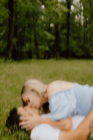 Couple sharing an intimate moment while lying in grass with trees in background, shot with dreamy blur effect.