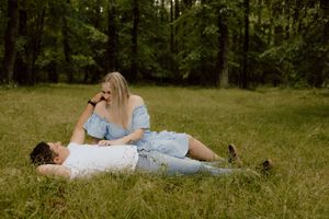 A couple in light blue attire laying together in a grassy meadow surrounded by tall trees on a sunny day.