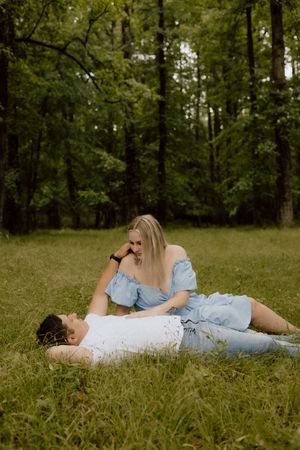 A couple in light blue outfits relaxes together on grass in a serene wooded setting during a summer day.