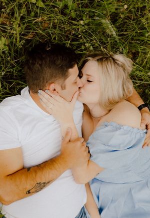 Romantic couple lying in grass sharing a tender kiss, wearing white and light blue clothing.
