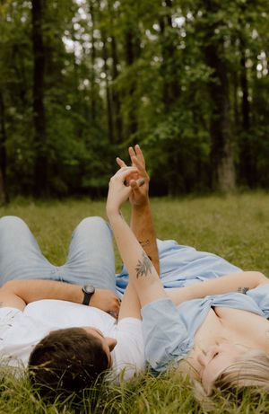 Two people lying on grass in a park making peace signs with their hands against a forest background.