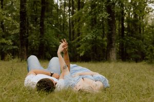 A couple lying in tall grass together in a peaceful wooded area during golden hour.