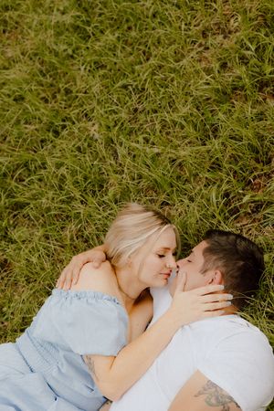 A couple laying together in green grass wearing light colored clothing share an intimate moment during a sunny outdoor photo session.