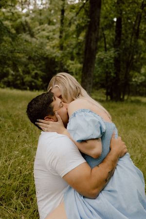 A couple embraces in a romantic kiss while standing in a lush green forest setting.