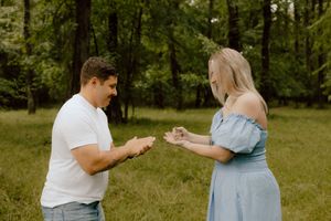 Two people standing in a green field exchange rings during what appears to be a proposal moment.
