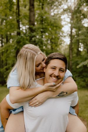 A loving couple shares a playful piggyback moment in a sunlit forest setting.