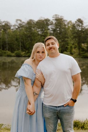 A couple stands together by a peaceful lake at sunset, with her wearing a light blue dress and him in a white t-shirt.