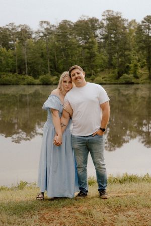 A couple poses together by a serene lake surrounded by trees, with the woman wearing a flowing light blue dress.