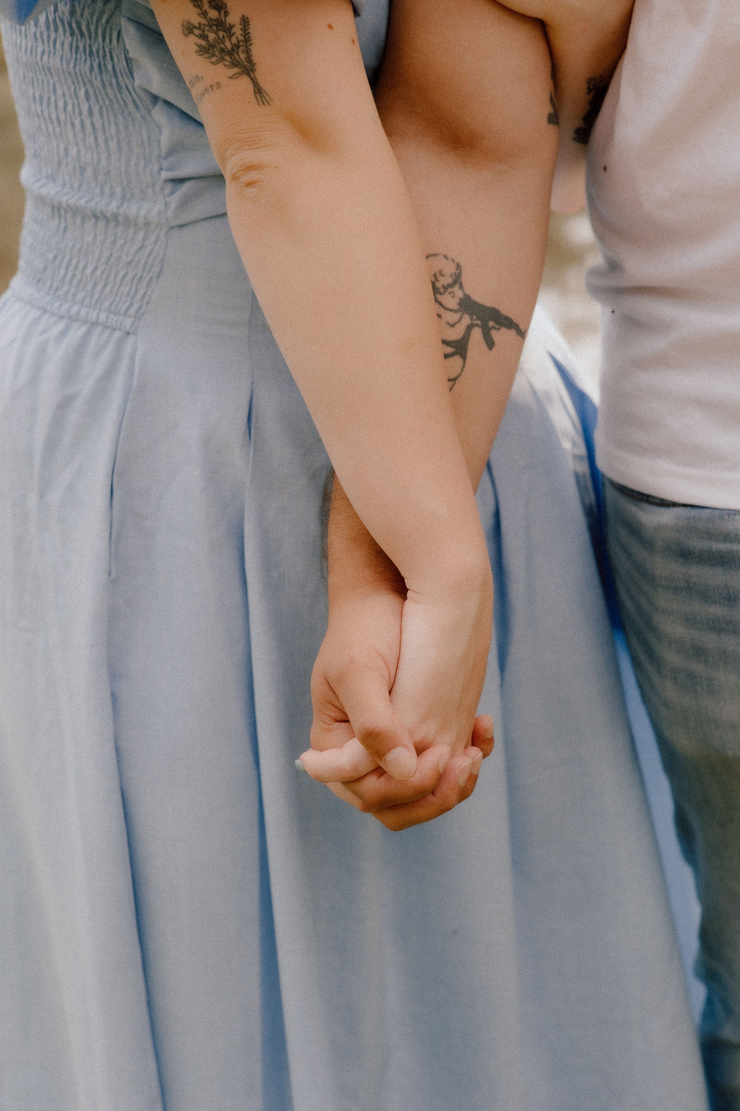 Close-up of clasped hands showing a tattoo detail against a light blue dress and casual attire.