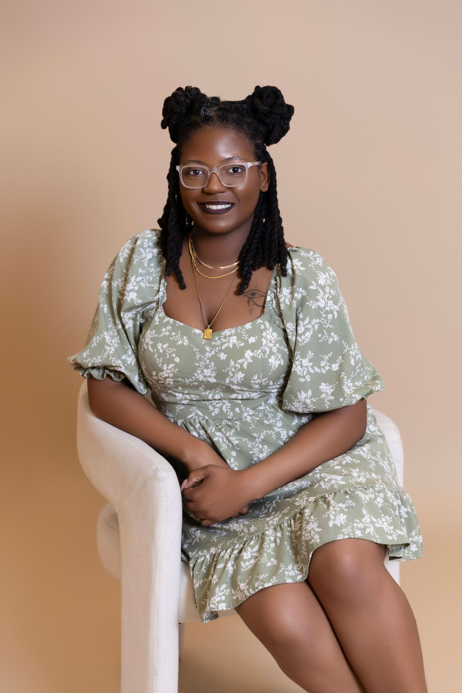A person in a floral dress sits on a white chair wearing glasses and space buns hairstyle.