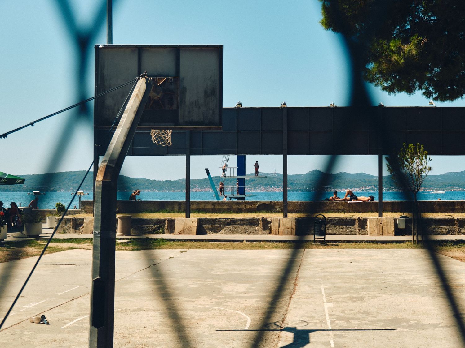 Basketball hoop on beachside court with ocean and boats visible in background through chain link fence.