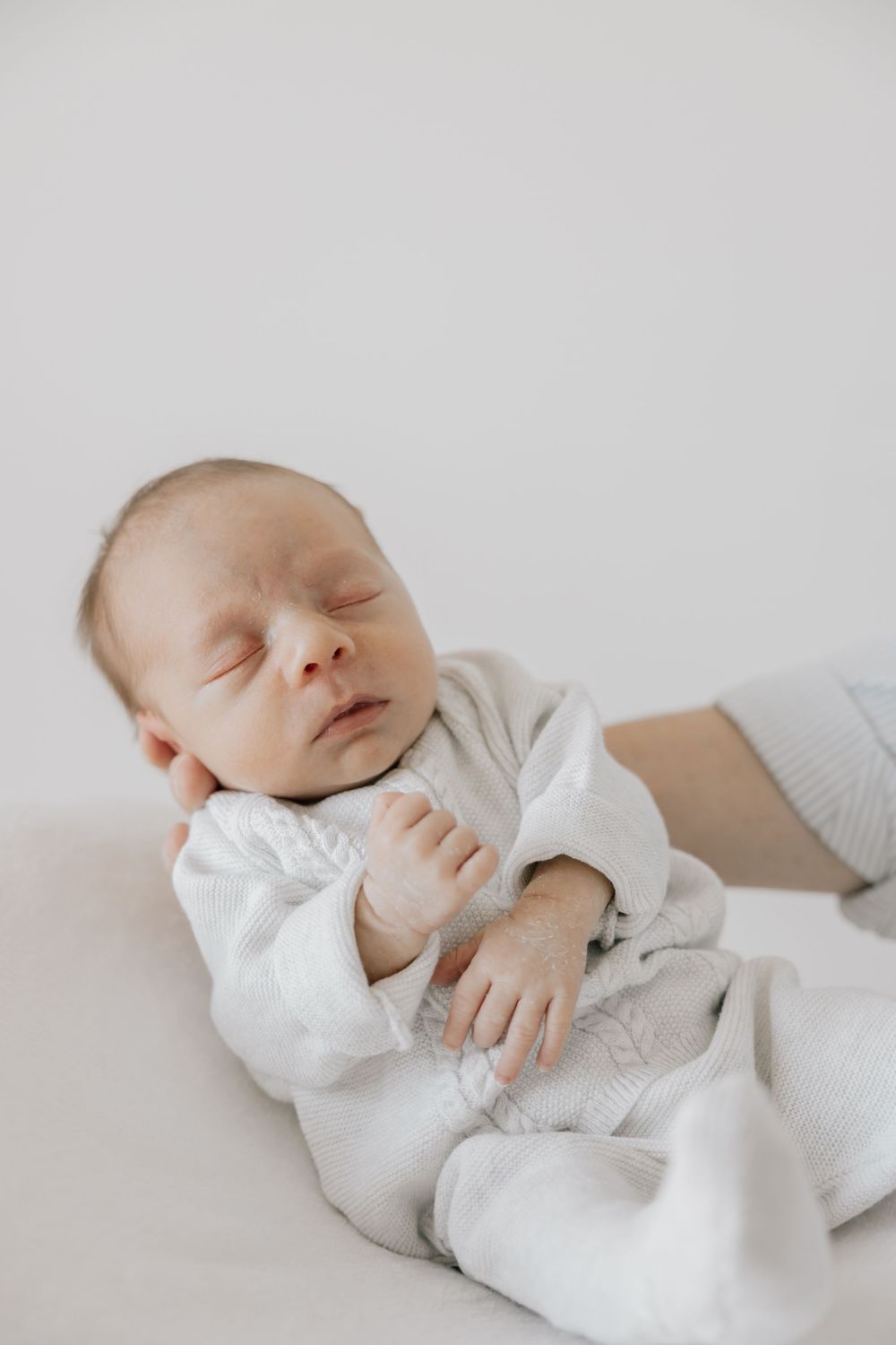 Peaceful newborn baby sleeping in white onesie, eyes closed, tiny hands folded on chest against soft white background.