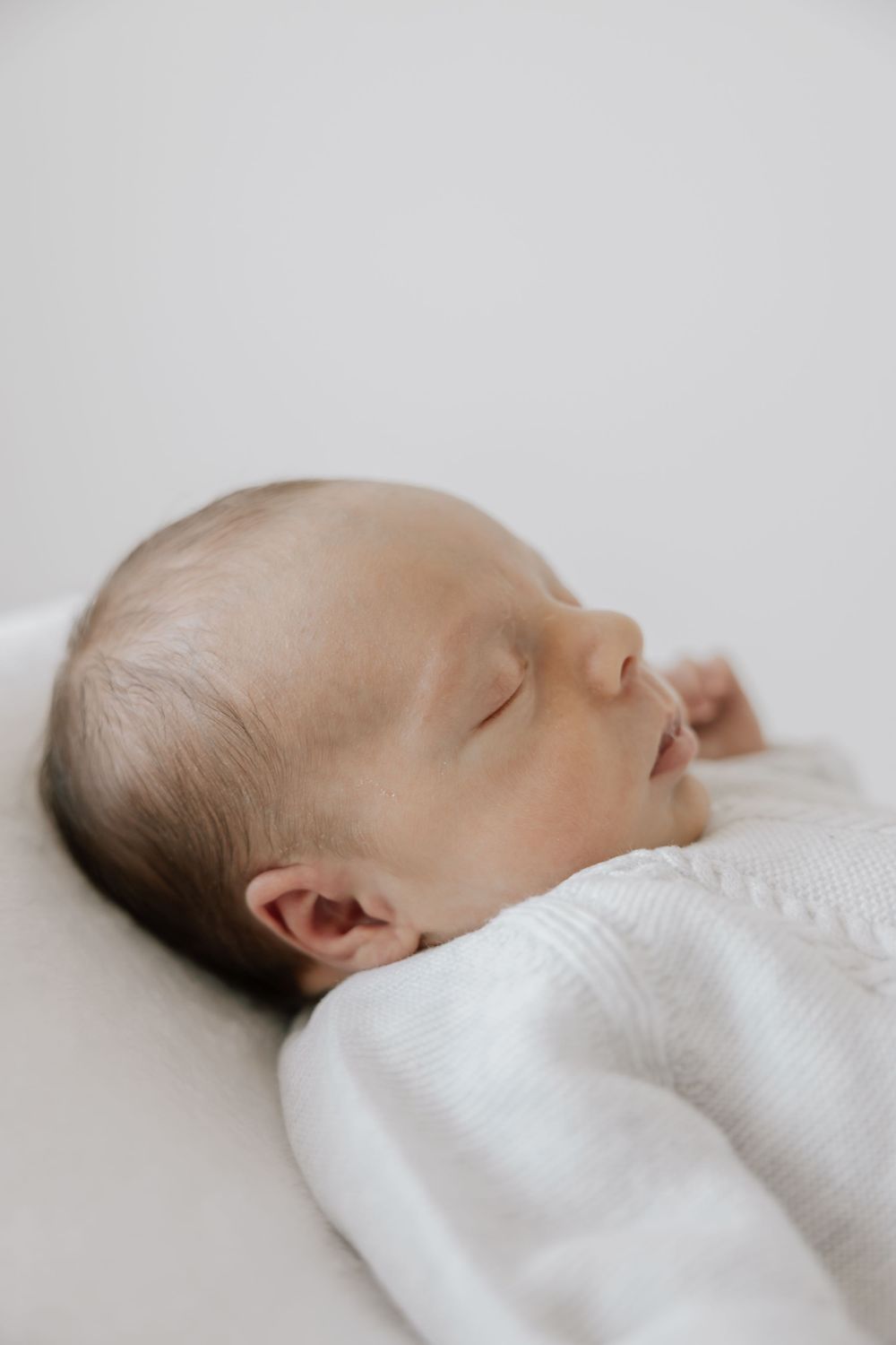 A peaceful newborn baby sleeping soundly, dressed in a white knit outfit against a soft white background.