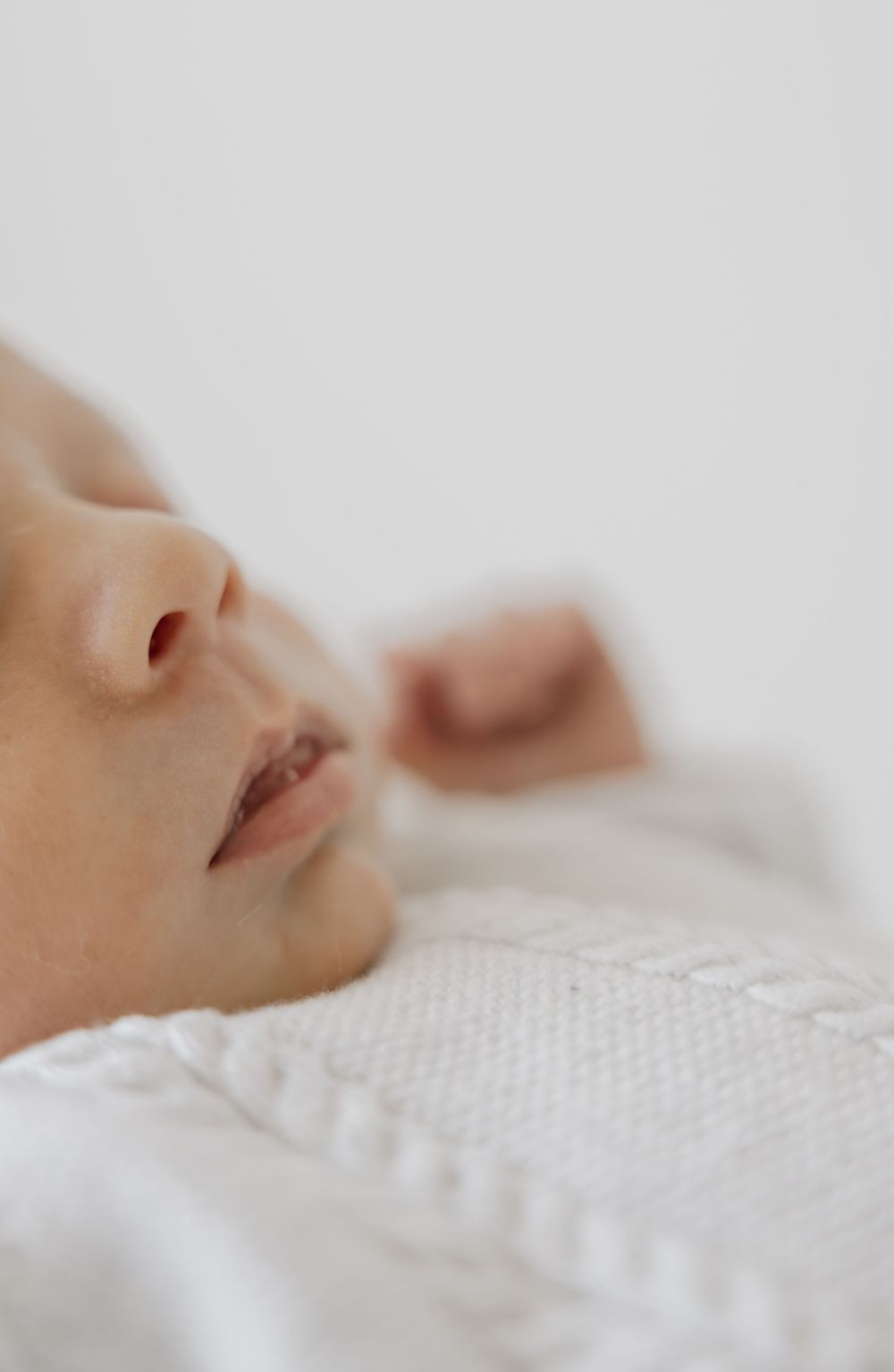 Peaceful newborn baby sleeping soundly, wrapped in a soft white knit blanket, close-up portrait photography.