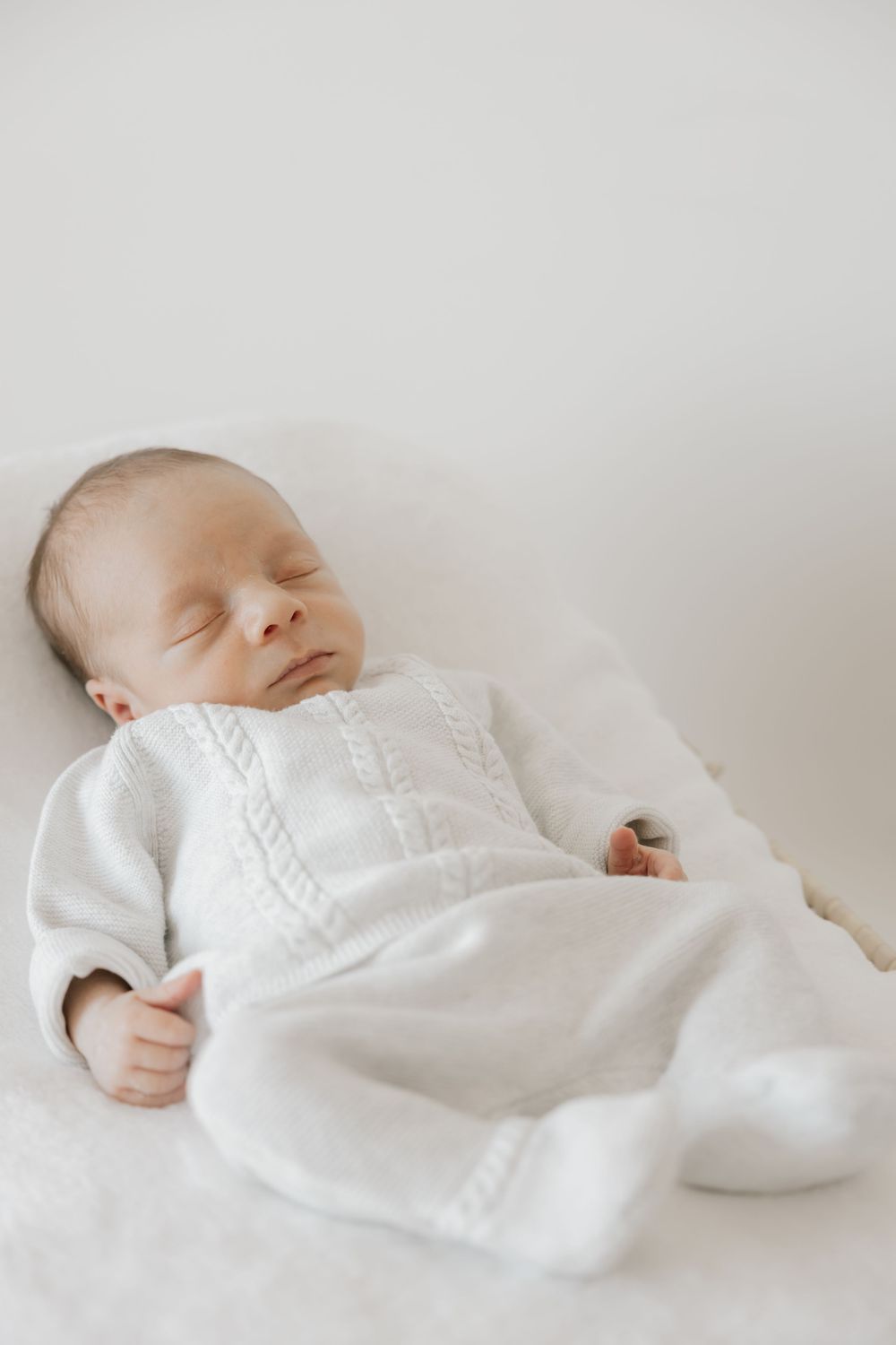 Peaceful newborn baby sleeping in white knit onesie on soft white blanket.