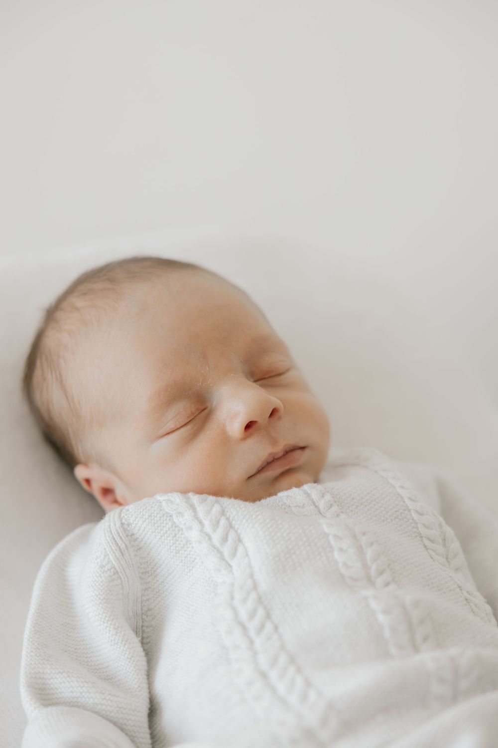 Peaceful newborn baby sleeping in white cable-knit outfit on soft white blanket.