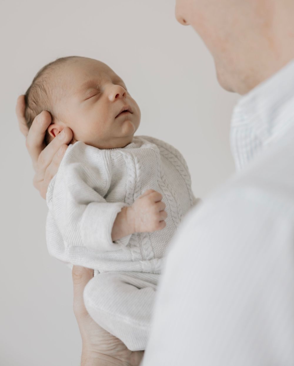 An adult gently cradling a sleeping newborn baby dressed in a white knit outfit against a soft white background.