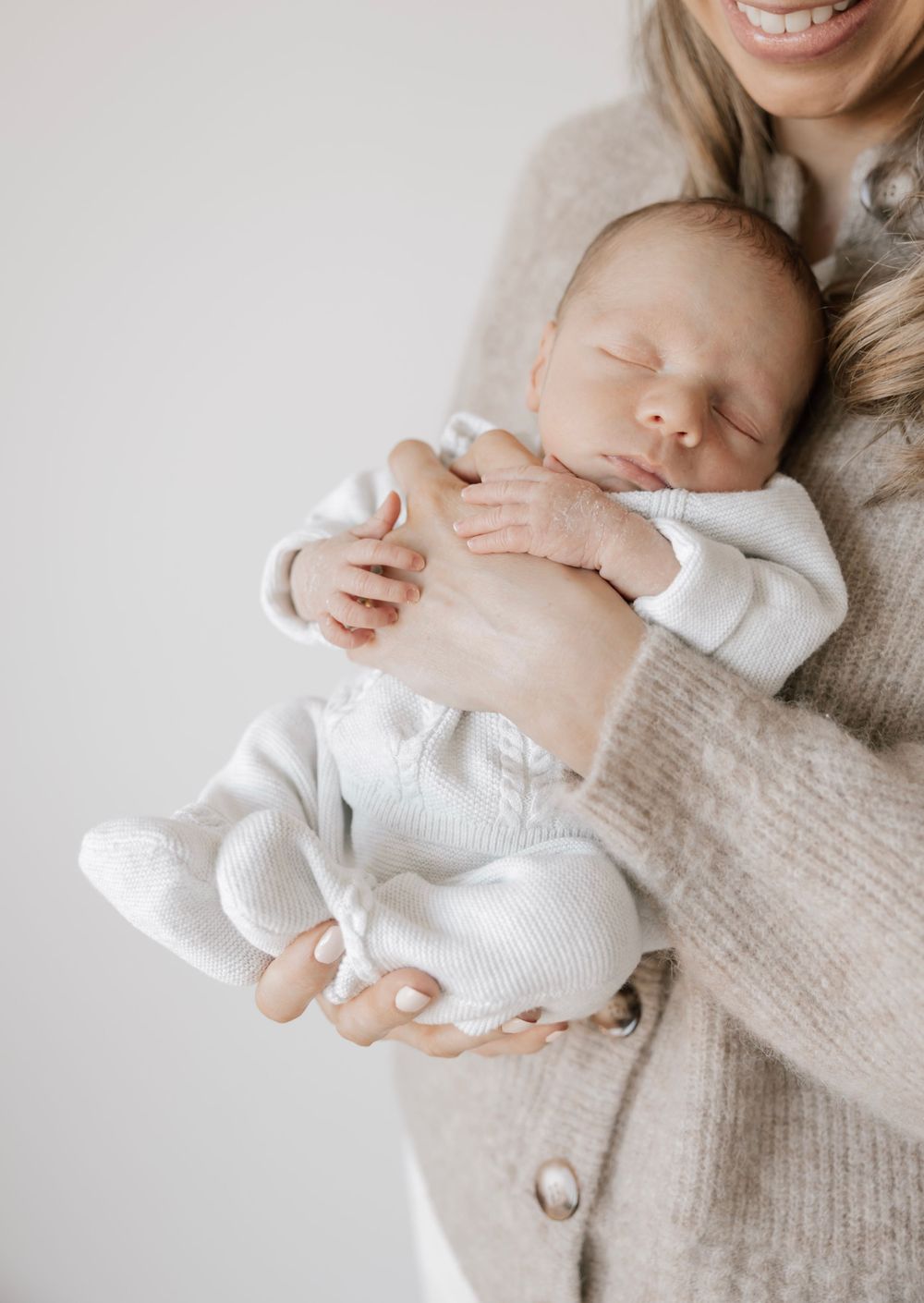 Smiling mother in beige cardigan cradling sleeping newborn baby dressed in white outfit against light background.