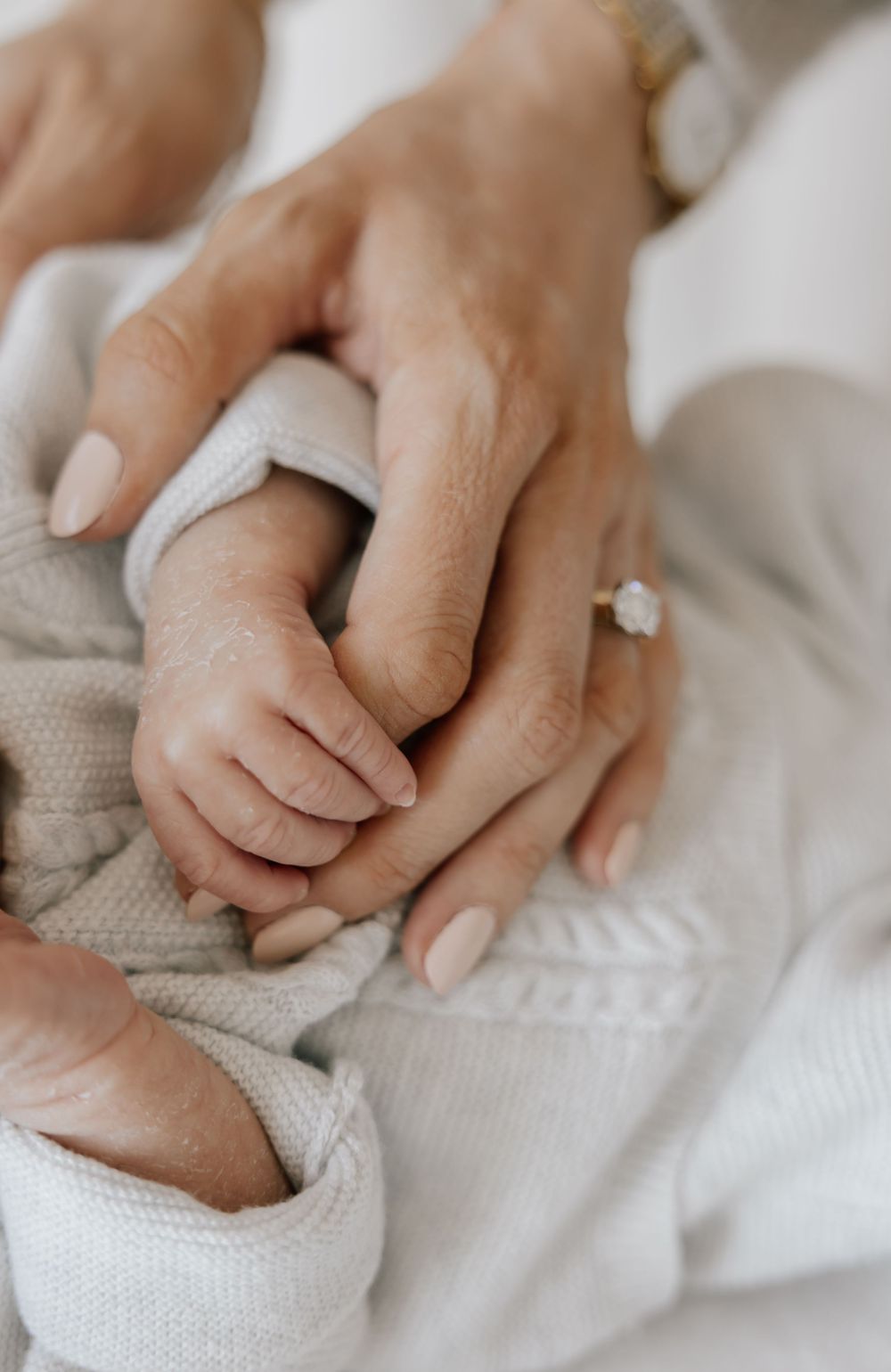 A mother's hand with a diamond ring gently holds her newborn baby's tiny fingers wrapped in a soft knit blanket.