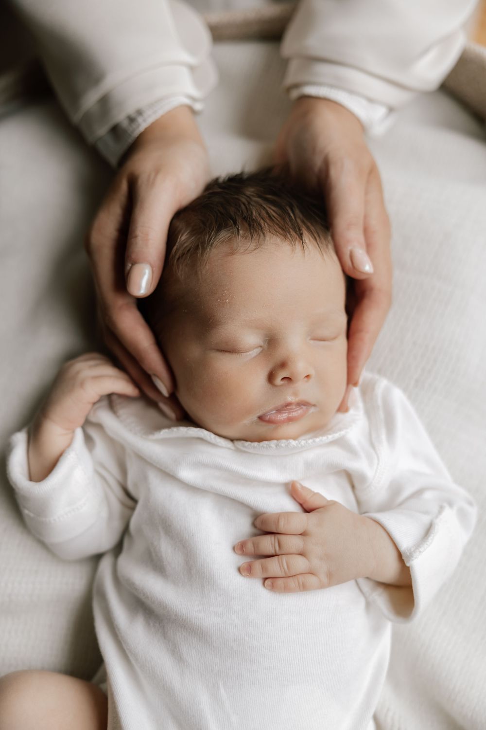 A sleeping newborn baby in white clothing with gentle hands cradling their head on white bedding.