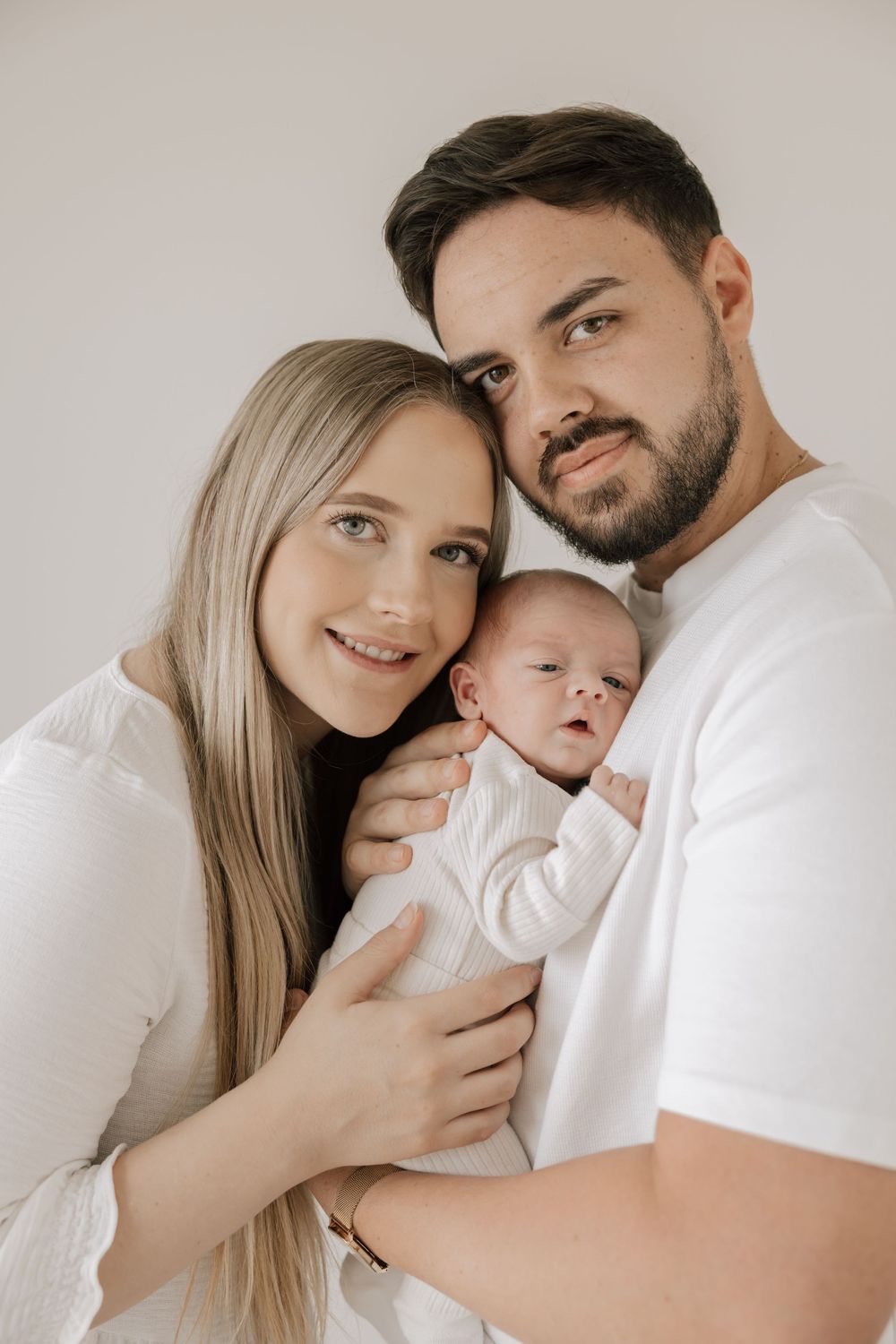 Young couple in white outfits smiling while tenderly holding their newborn baby against a light background.