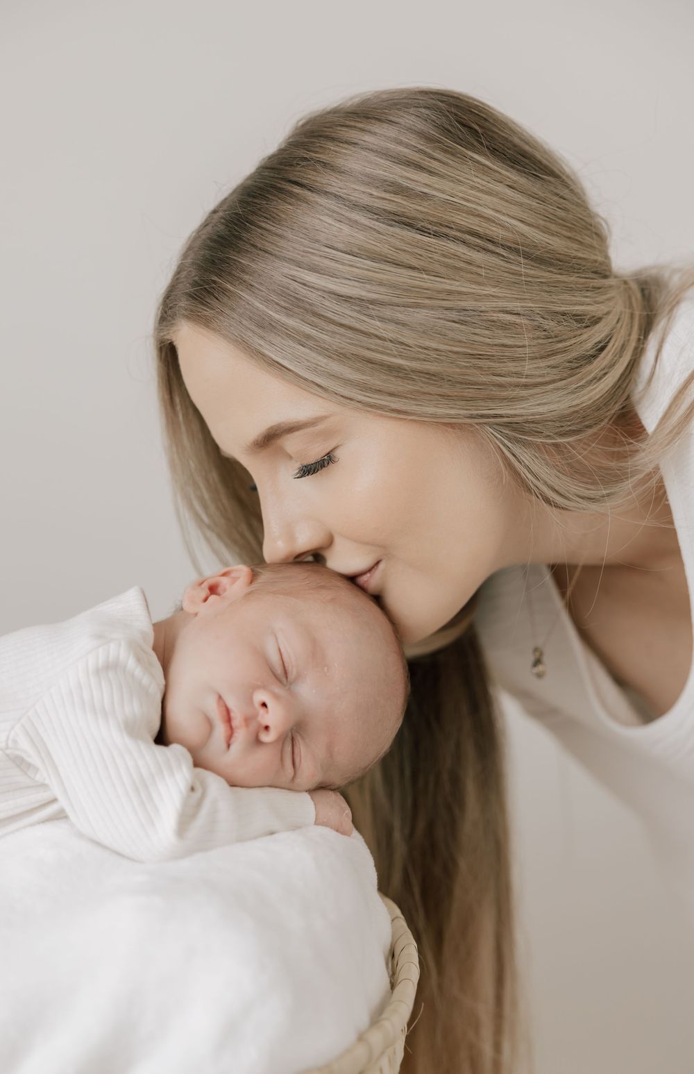 A young mother with long blonde hair gently kisses her sleeping newborn baby wrapped in a white swaddle.