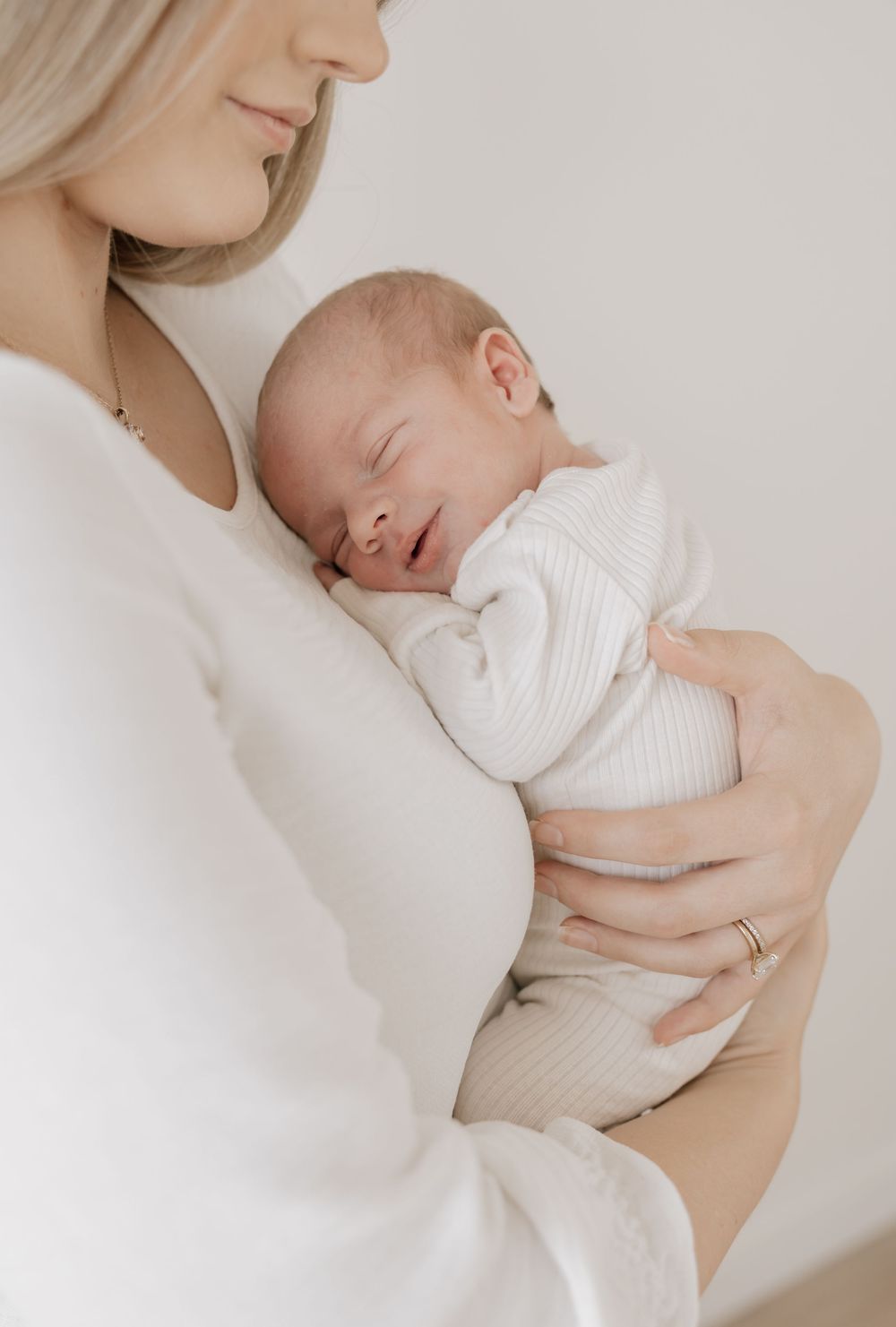 A mother in white gently cradles her sleeping newborn baby, both dressed in soft white clothing.