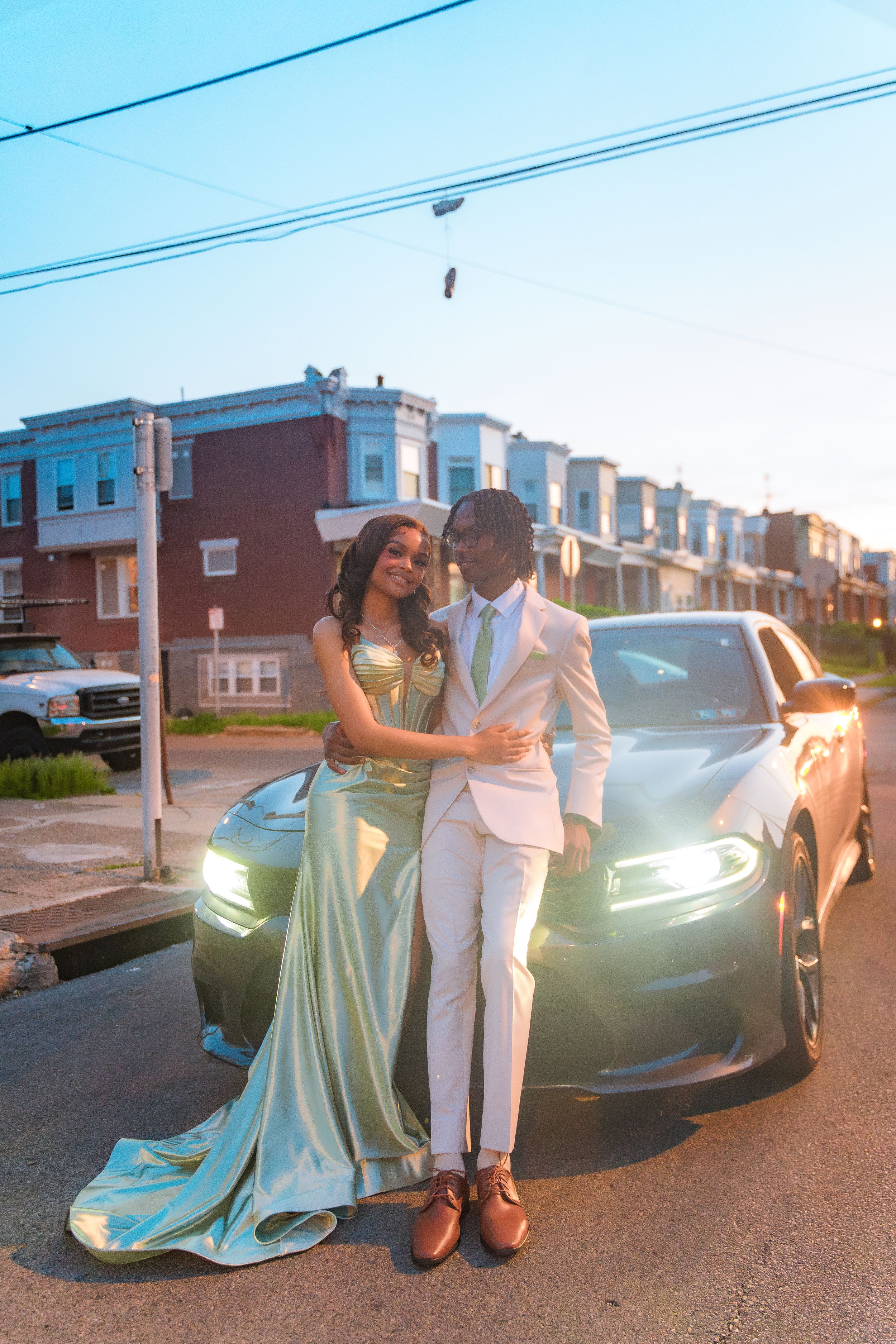 Couple in elegant formal wear standing beside car on street at dusk.