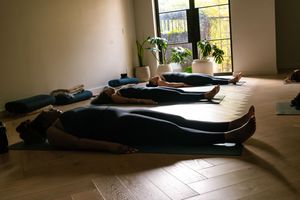 People lie on black yoga mats during a meditation or relaxation session in a sunlit studio space.