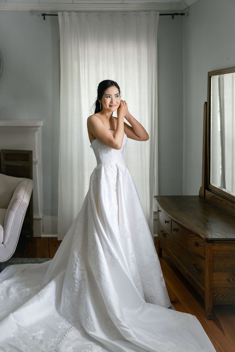A bride wearing a classic white ball gown poses elegantly against a window with sheer curtains.