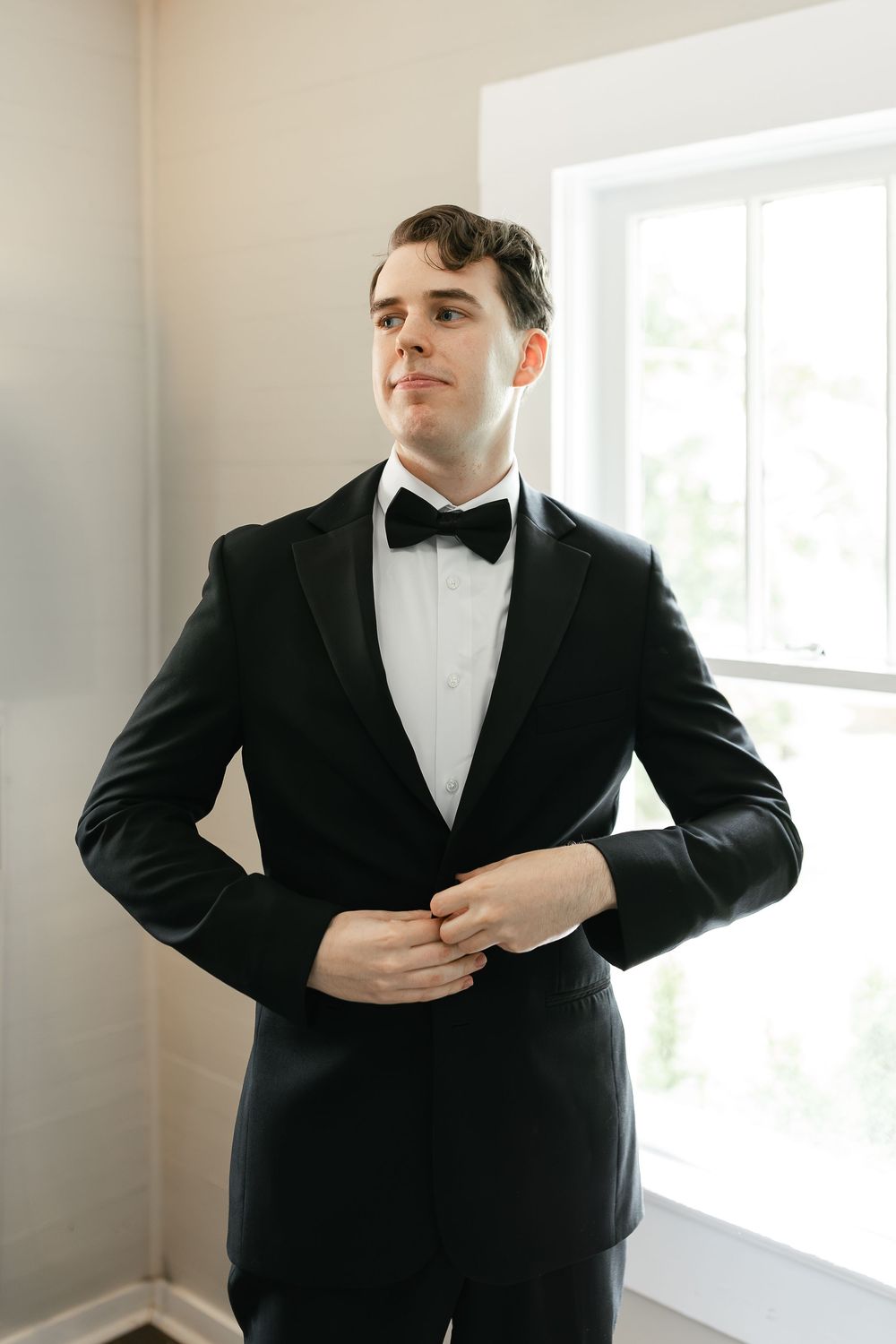 A person in a classic black tuxedo and bowtie adjusts their cuff in front of a bright window.