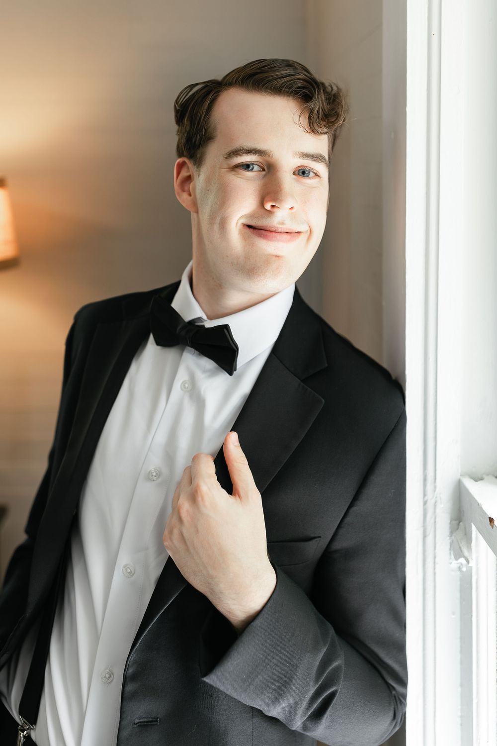A groom in classic black tuxedo and bowtie poses by a window.