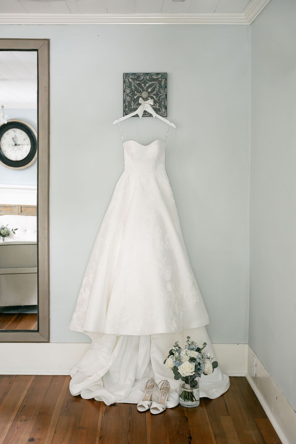 A classic white wedding dress with draped fabric hangs elegantly against a light blue wall with hardwood flooring below.