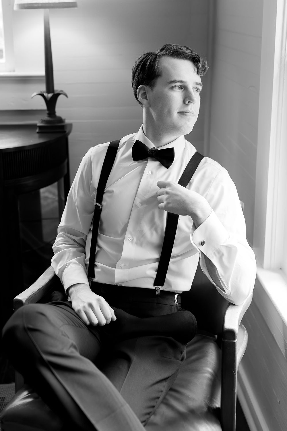 Young ring bearer in white shirt and black suspenders sitting thoughtfully by window.