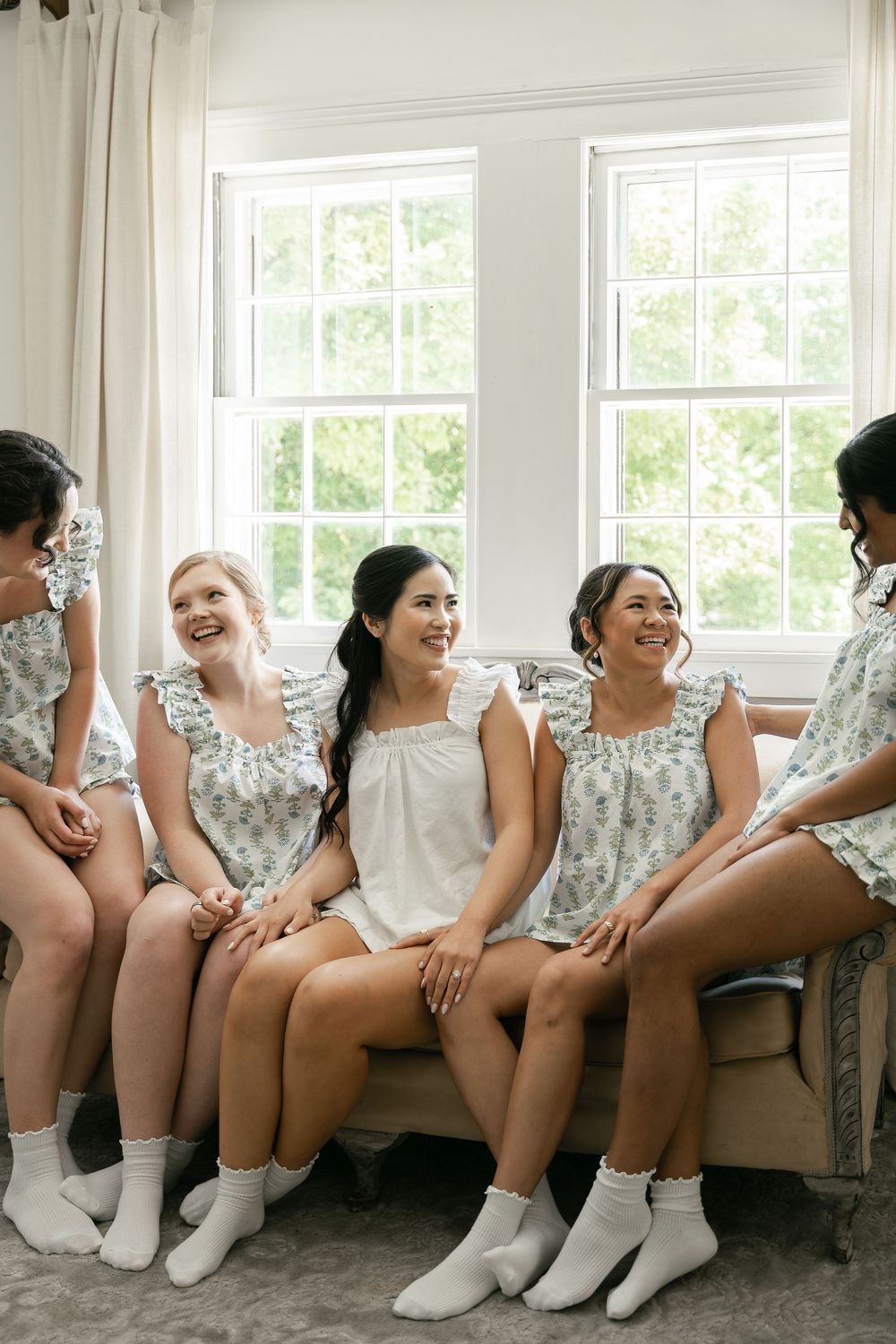 A bridal party in floral robes and white socks sits together on a couch sharing a candid moment.