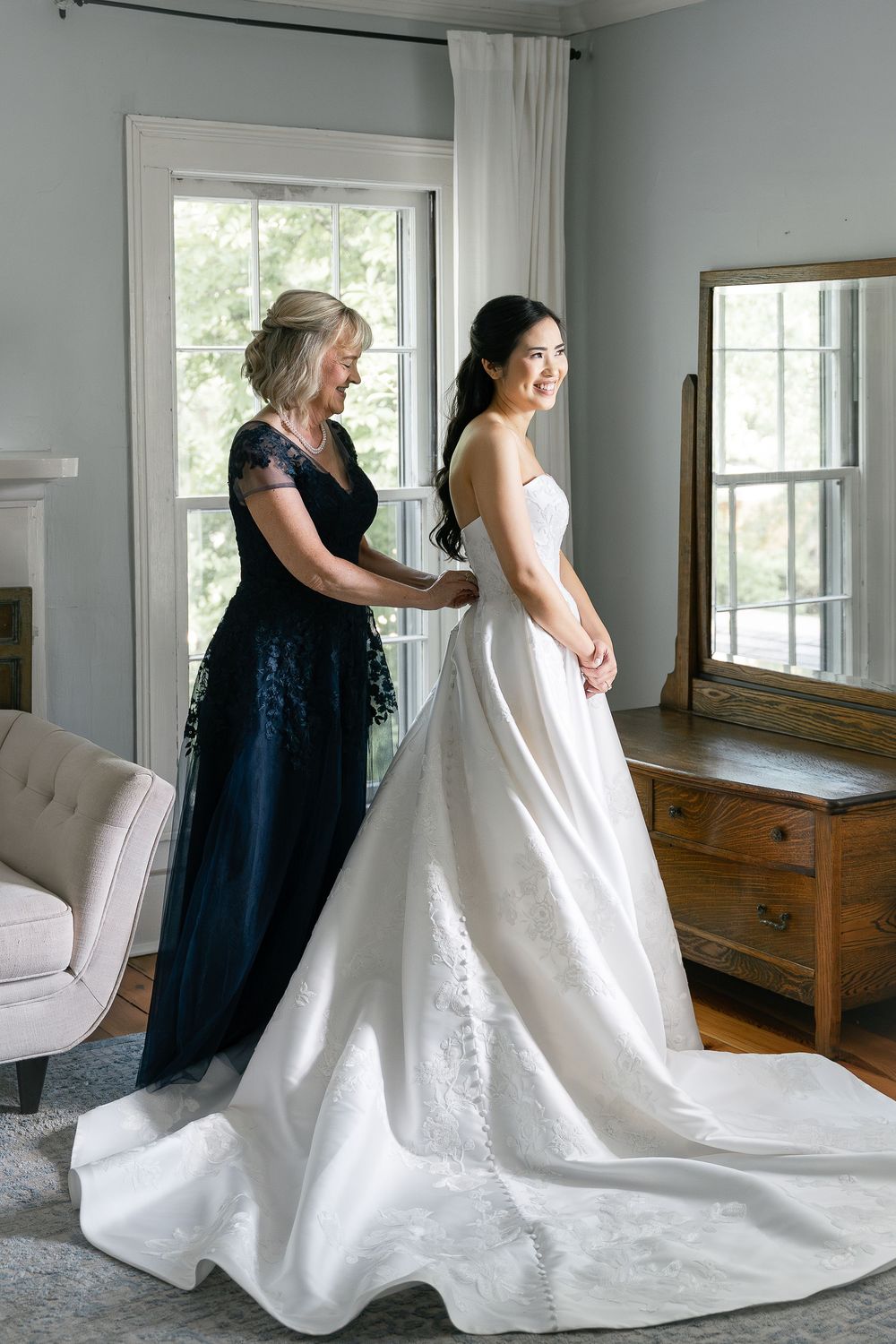 Wedding dress being adjusted and styled in a bright, window-lit dressing room.