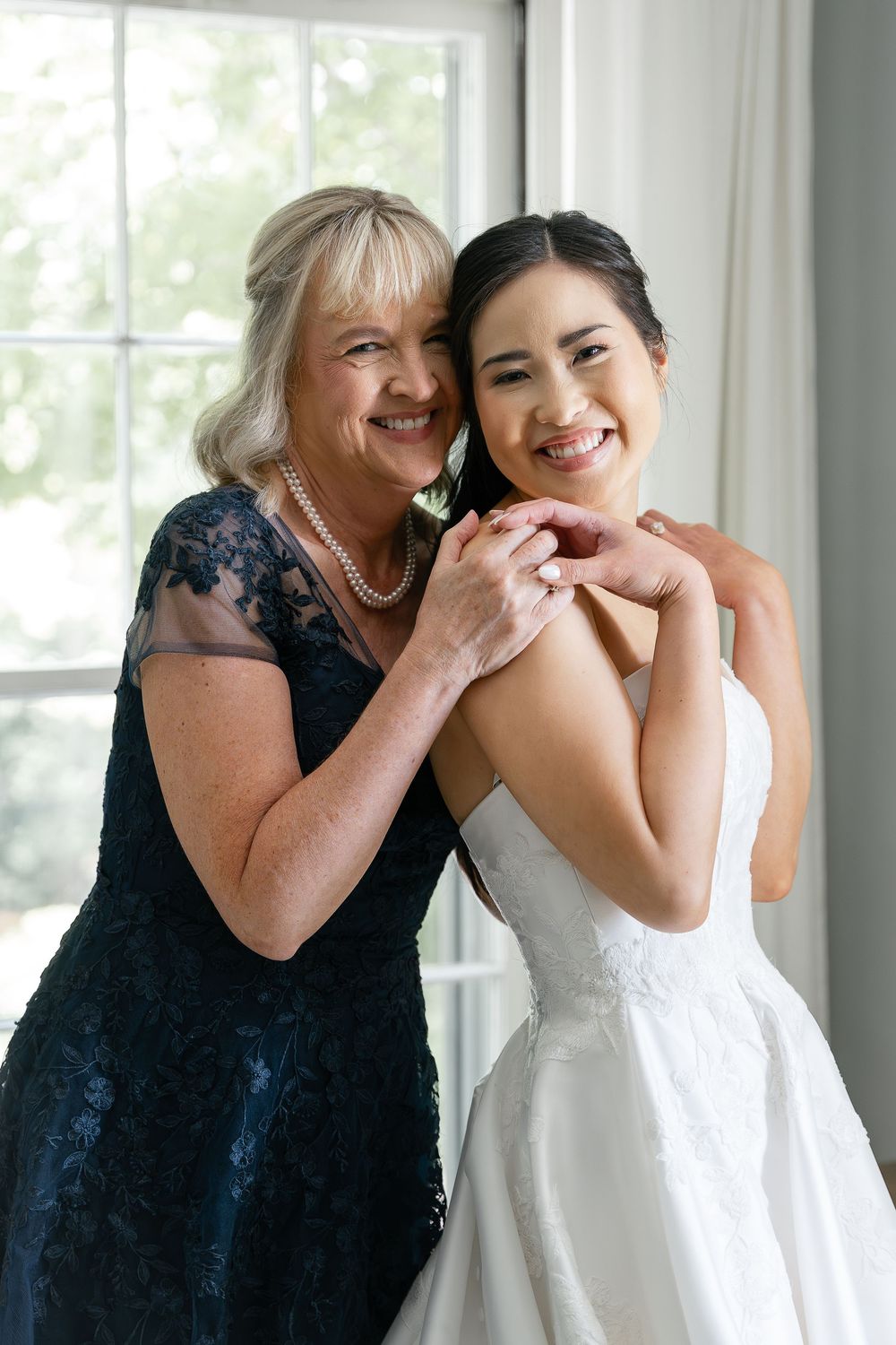 Two smiling women share a heartwarming embrace by a window, one wearing a navy lace dress and one in a white wedding gown.