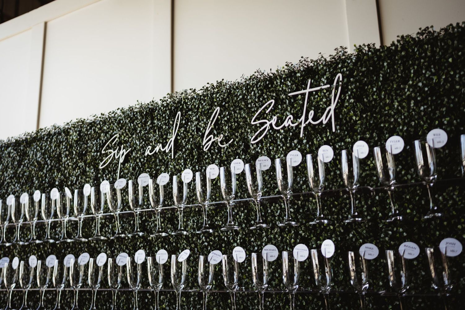 Rows of champagne glasses lined up against a dark wall with green foliage and illuminated 'Sip and be Seated' neon sign.