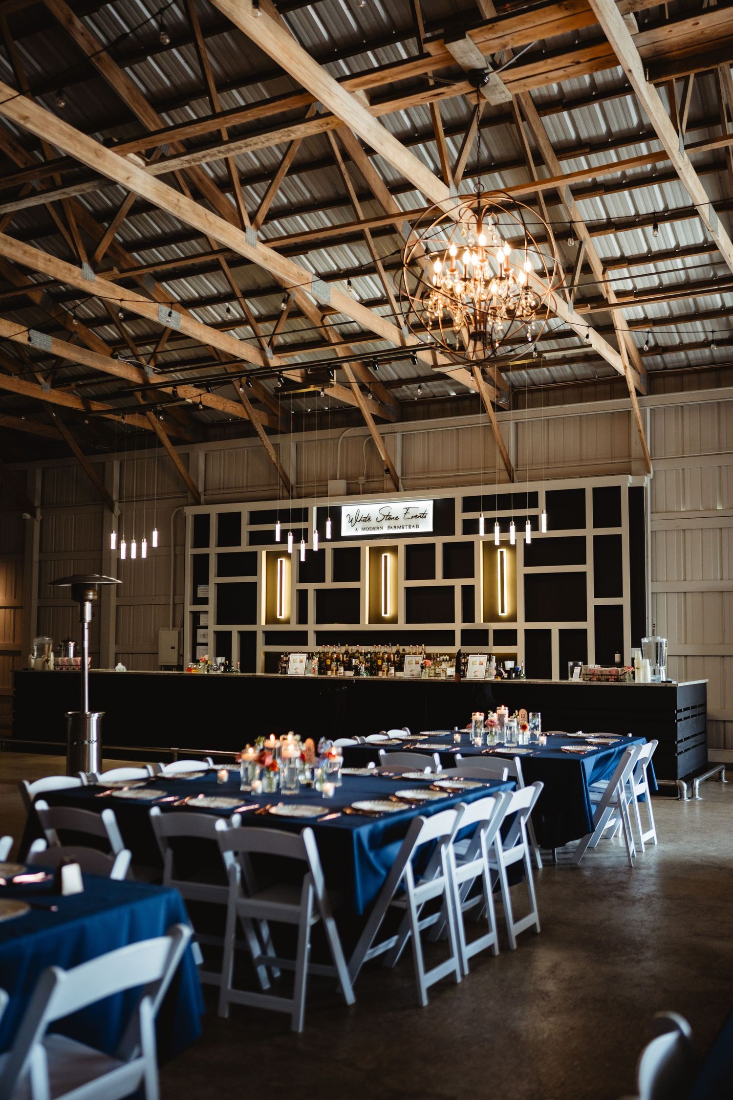 An elegant event space with exposed beams, crystal chandelier, and navy blue dining tables arranged for a formal gathering.