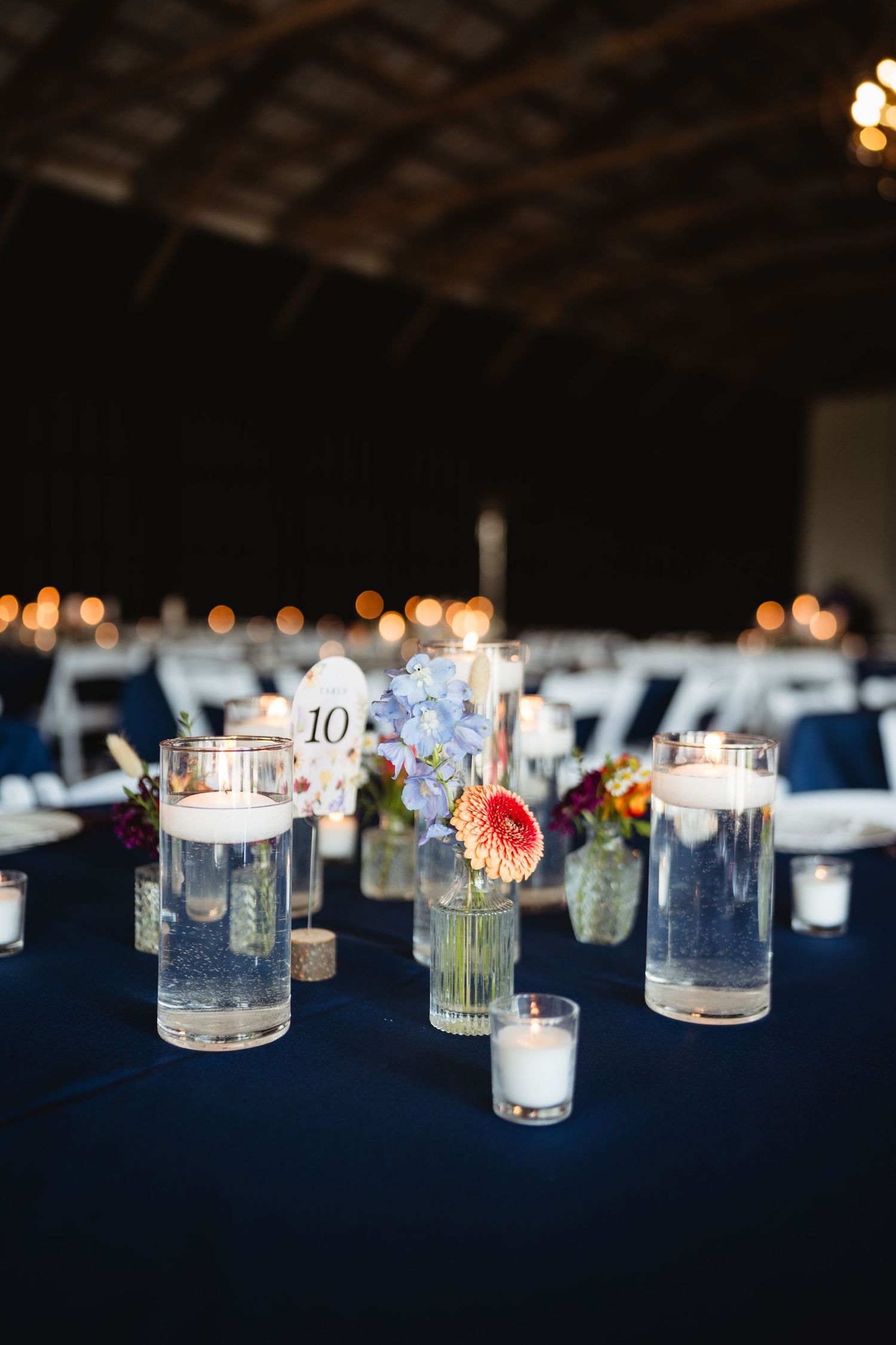 Wedding reception table decorated with floating candles, blue hydrangeas and table number on dark navy tablecloth.