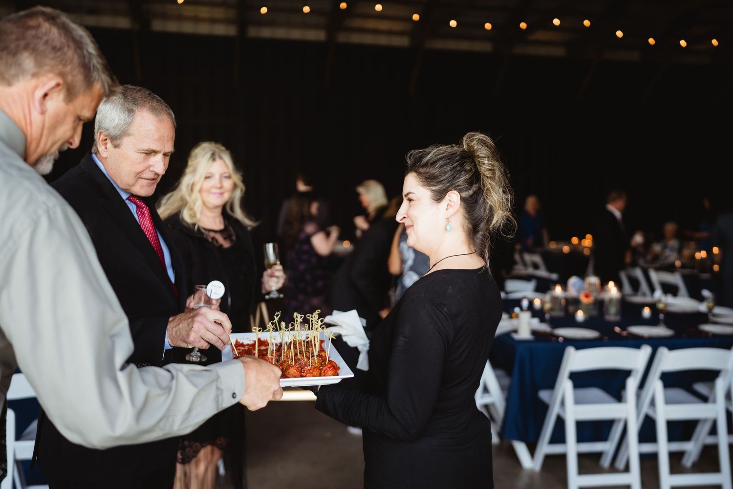 Guests being served appetizers at an evening event with string lights and white folding chairs in the background.