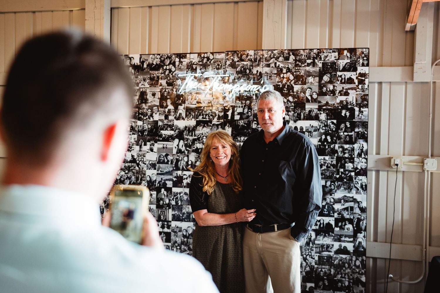 Photographer takes picture of couple posing together against black and white photo collage wall.