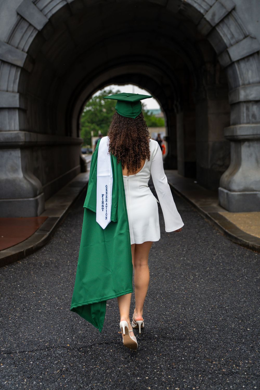 Graduate walks through stone archway wearing white dress and green graduation regalia.