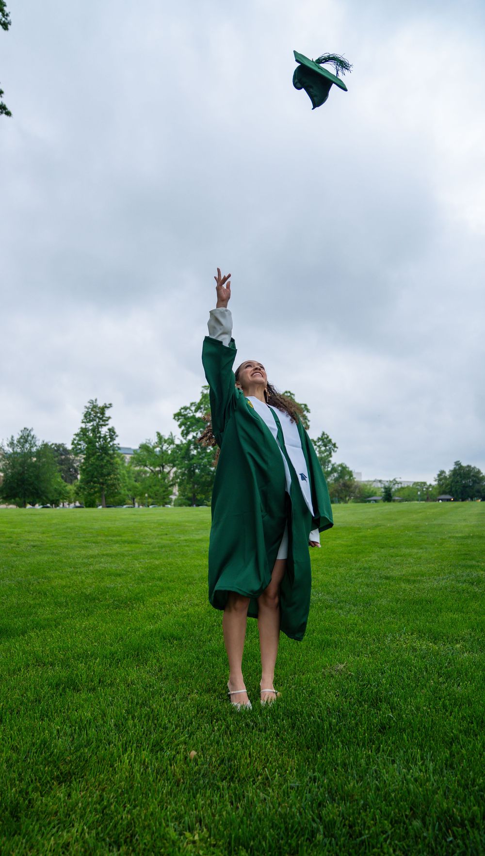Graduate throws cap in celebration on green lawn under cloudy sky.